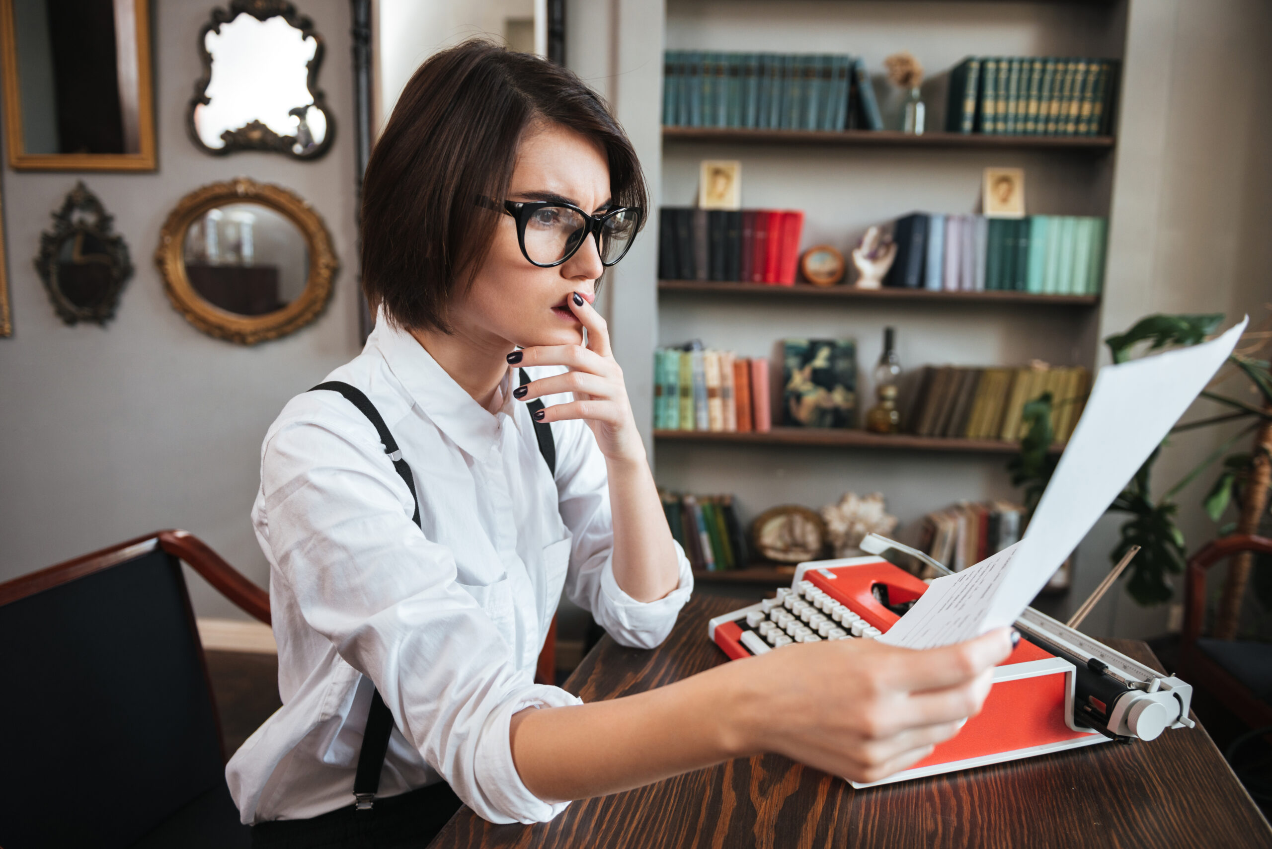 Side view of thoughtful authoress in glasses and white shirt sitting by the table with paper and typewriter